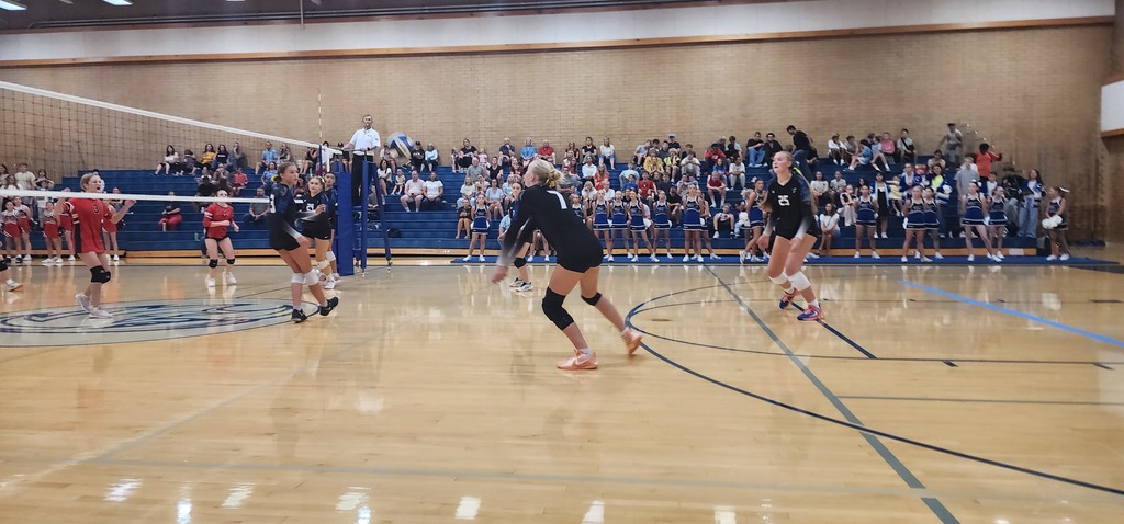 Fairfield's number 7 is passing a ball while the other 4 girls are setting up to get the ball. On the left side of the pic, behind the net is South Davis' team. The ref is on a stand in the middle. In the background is the crowd in the bleachers.