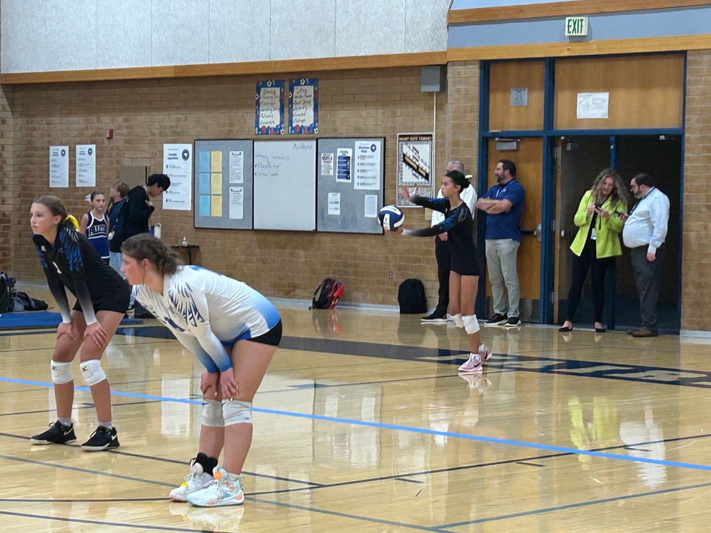 A Fairfield player is serving the ball. Two of Fairfield's players, one in a white shirt and the other in a black shirt are bent at the waist watching the team out of the picture to the left. In the background are several people standing next to a wall.