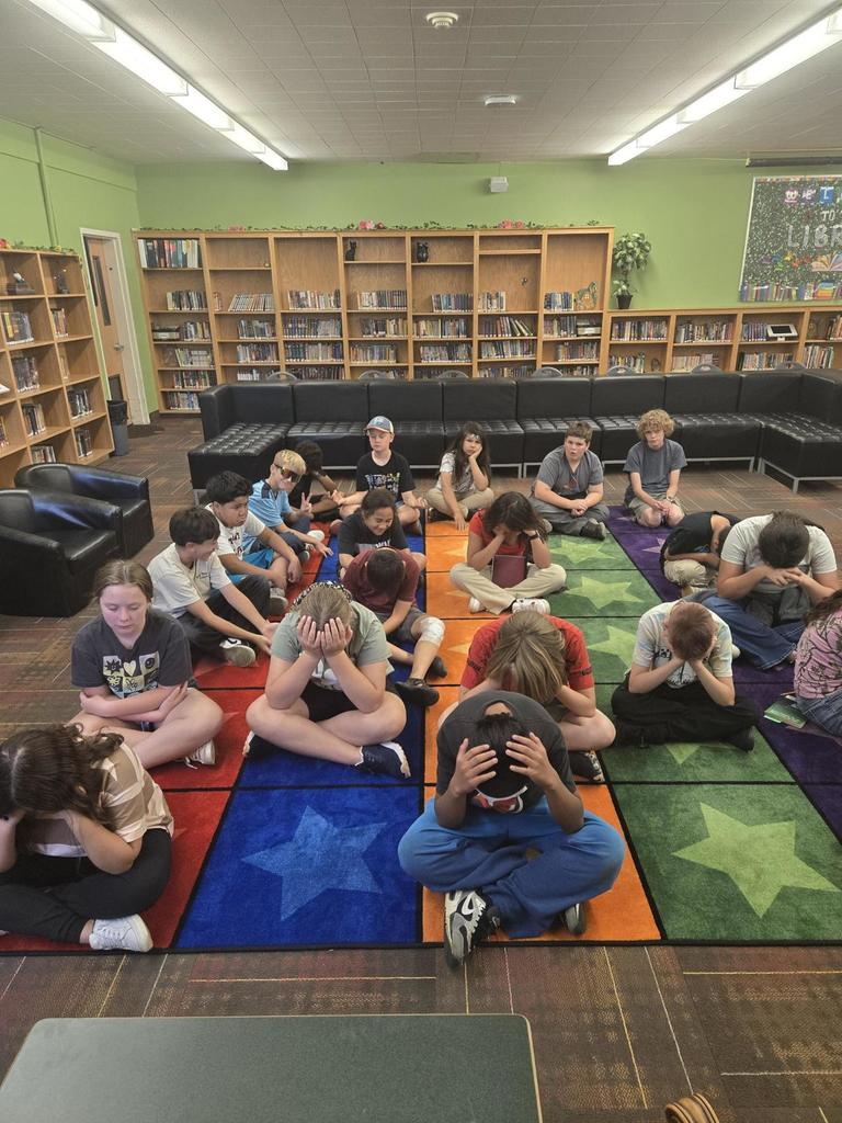 a group of 6th grade students sitting on a colorful rug who have the heads down, meditating. 
