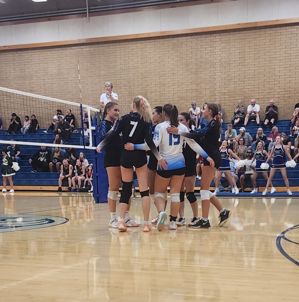 The Varsity team is huddled up on the court. The ref is standing above the net, and in the background are fans seated in the bleachers with cheerleaders standing in front of them.