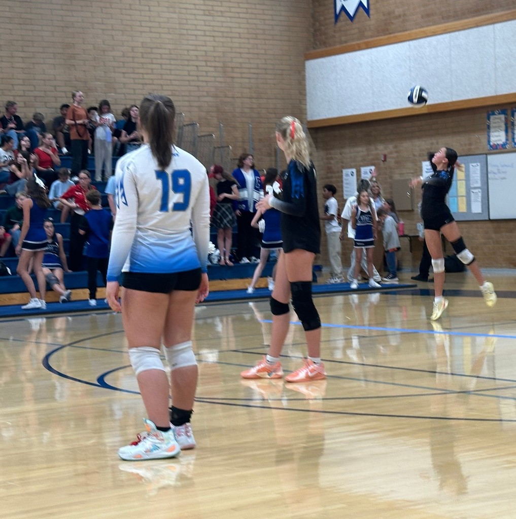 One of Fairfield's girls is serving the ball from the back while two players, one in a white jersey and the other in a black jersey are looking back at the server. The crowd is in the background seated in the bleachers.