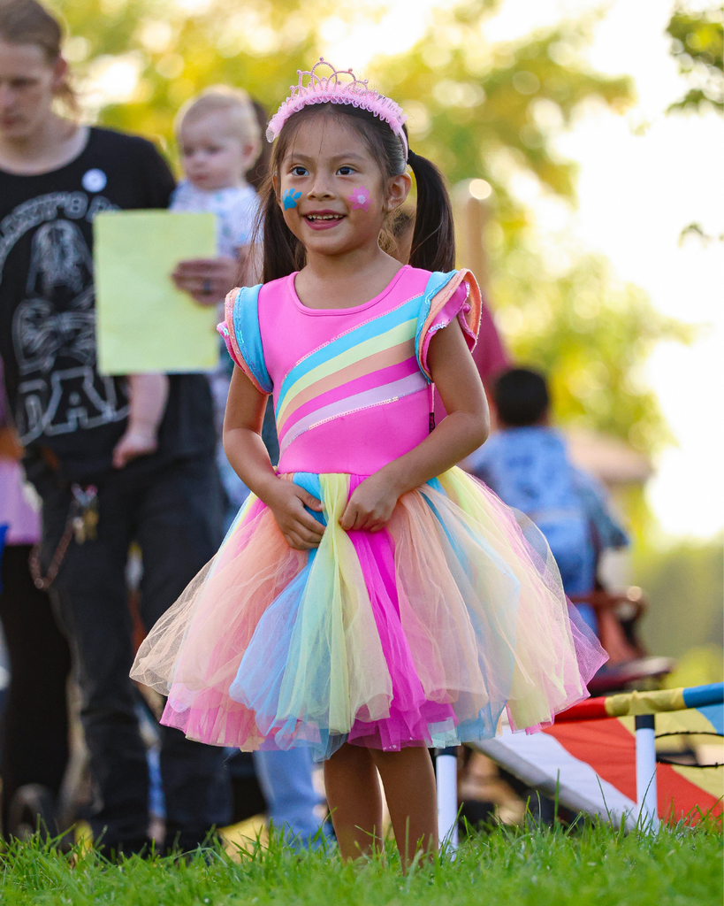 A girl in a rainbow tutu with her face painted smiles to the distance.