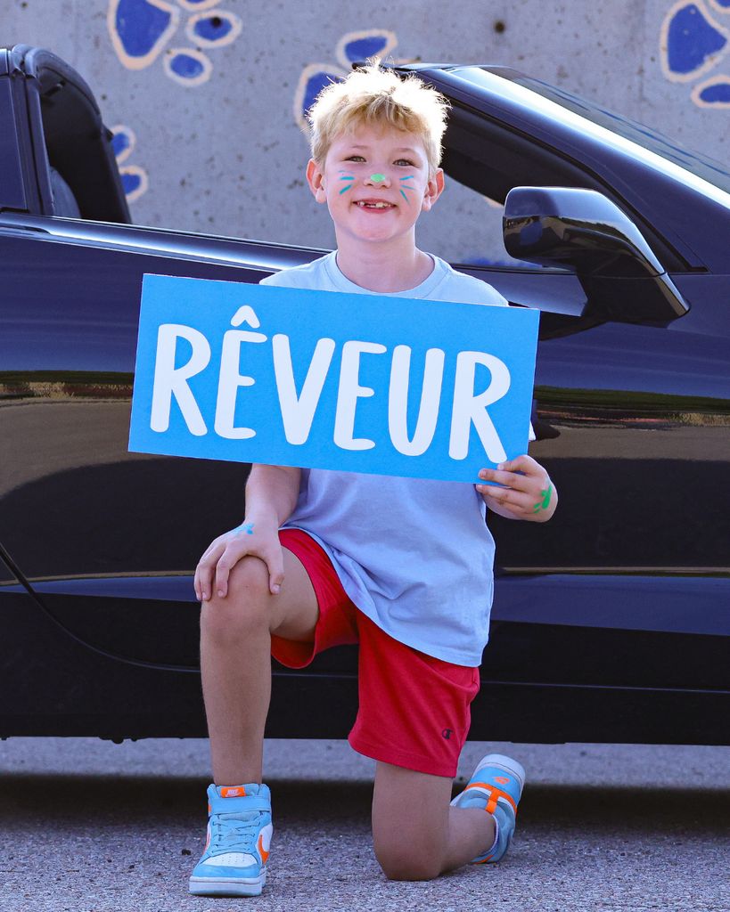 A student kneeling in front of a car with his face painted holds a sign that reads "rêveur."