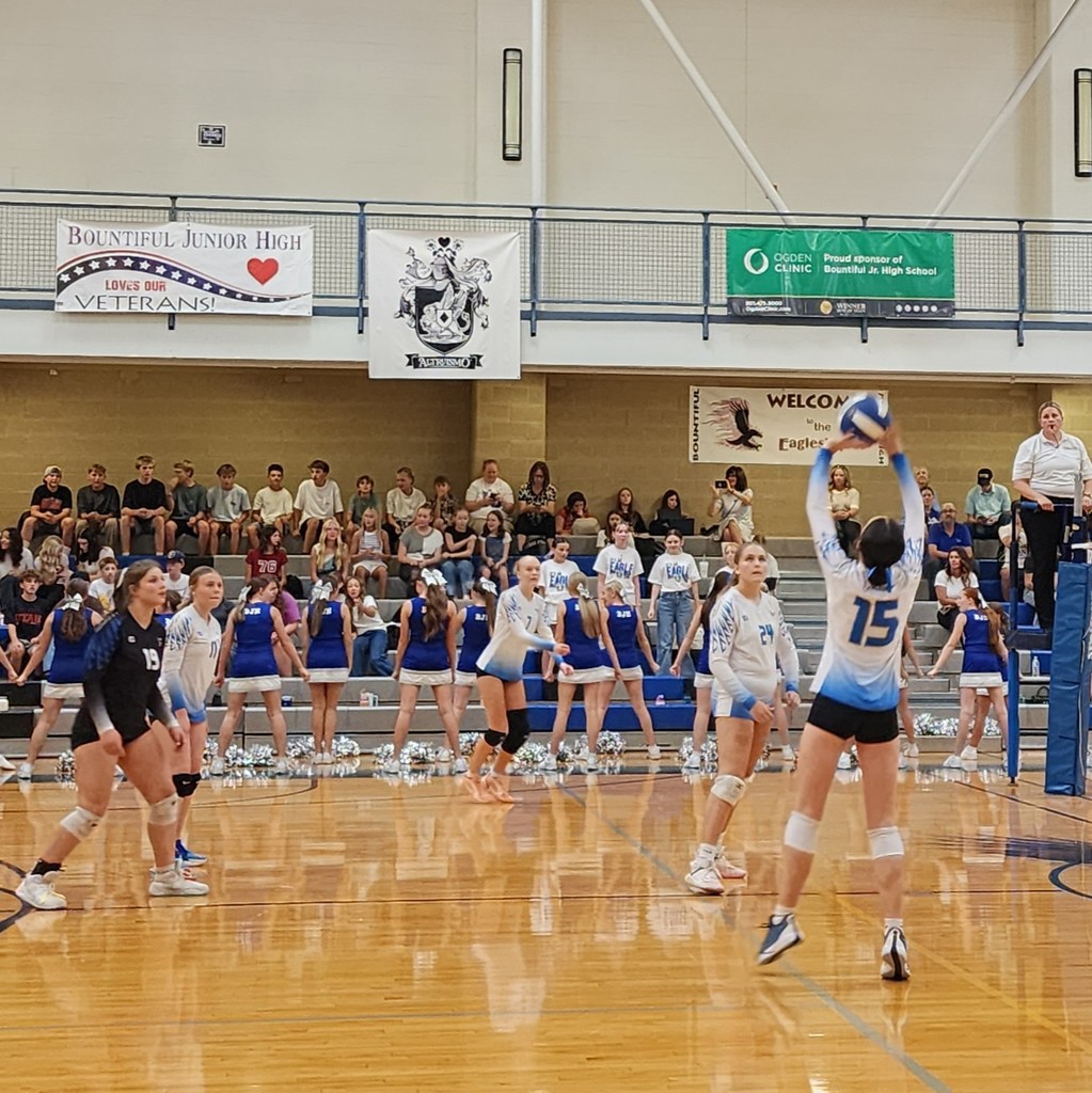 A Fairfield setter is setting the hitters towards the back.  Four players are watching the setter.   The ref is standing at the net and the crowd is seated in the background.  Bountiful's cheer squad is standing between the court and the bleachers.