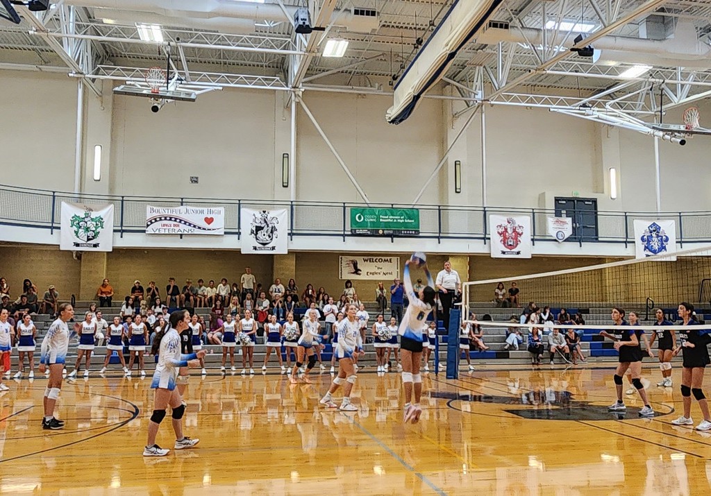 Fairfield's setter is jump setting the ball towards a hitter in the background.  The remaining girls are watching and waiting to move.  There is a ref on a stand at the net and there is  a crowd in bleachers in the background.