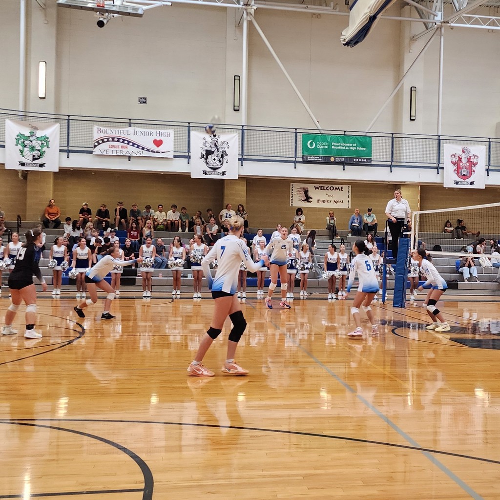 A girl on Fairfield's volleyball team is passing the ball.  The other girls are setting up to hit the ball over.  There is a ref standing at the net, and a crowd seated in the bleachers in the background.