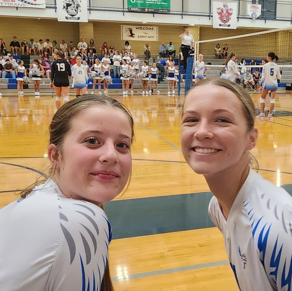 Two of Fairfield's JV players are smiling at the camera in the foreground.  Behind them, the Varsity team is standing, waiting for a serve to occur.  The Ref is standing at the net.  The crowd is in the background, with Bountiful's cheer squad in standing between the court and the bleachers.