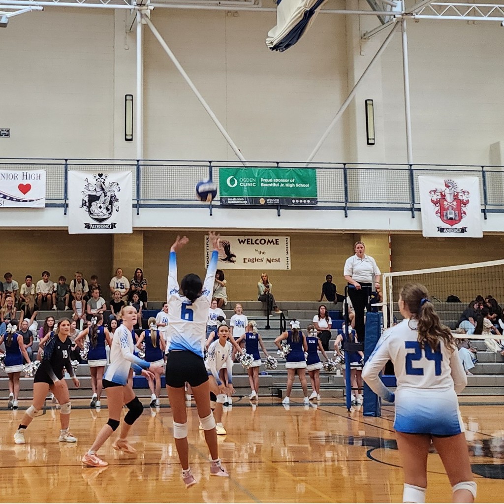 Fairfield is setting a ball to the hitters towards the b ack.  The libero on the left and the other player in the foreground are watching the play.  There is a ref on a stand at the net and the crowd is seated in bleachers in the background.  Bountiful's cheer squad is in front of the bleachers cheering.