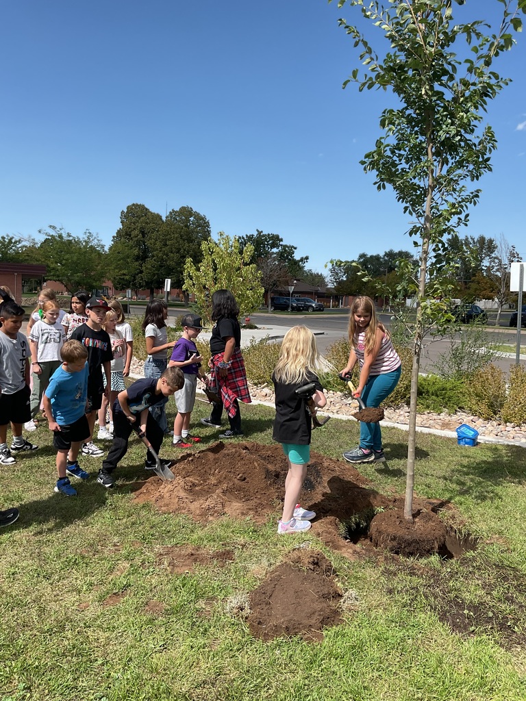 Students holding shovels full of dirt next to a large hole with a thin tree placed inside. They are helping to cover the roots of the newly planted tree