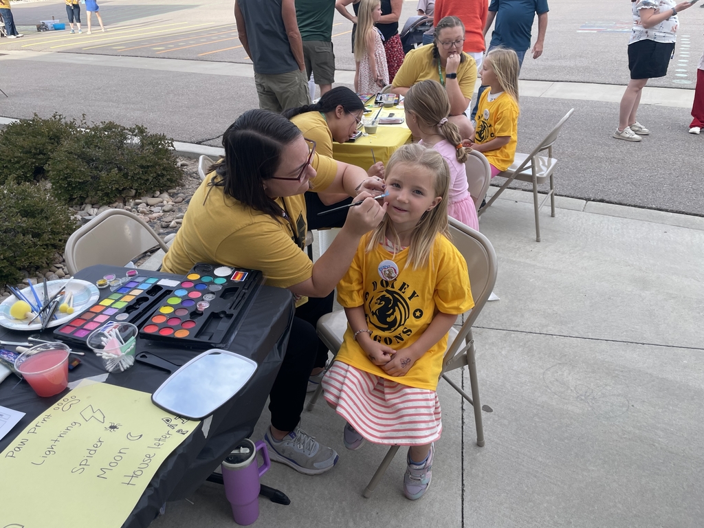 Students sitting on chairs outside while staff paints their face with festive images.