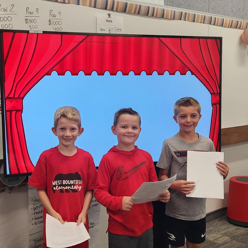 Three smiling boys holding papers and standing in front of a screen that has curtains like the boys are on a stage.