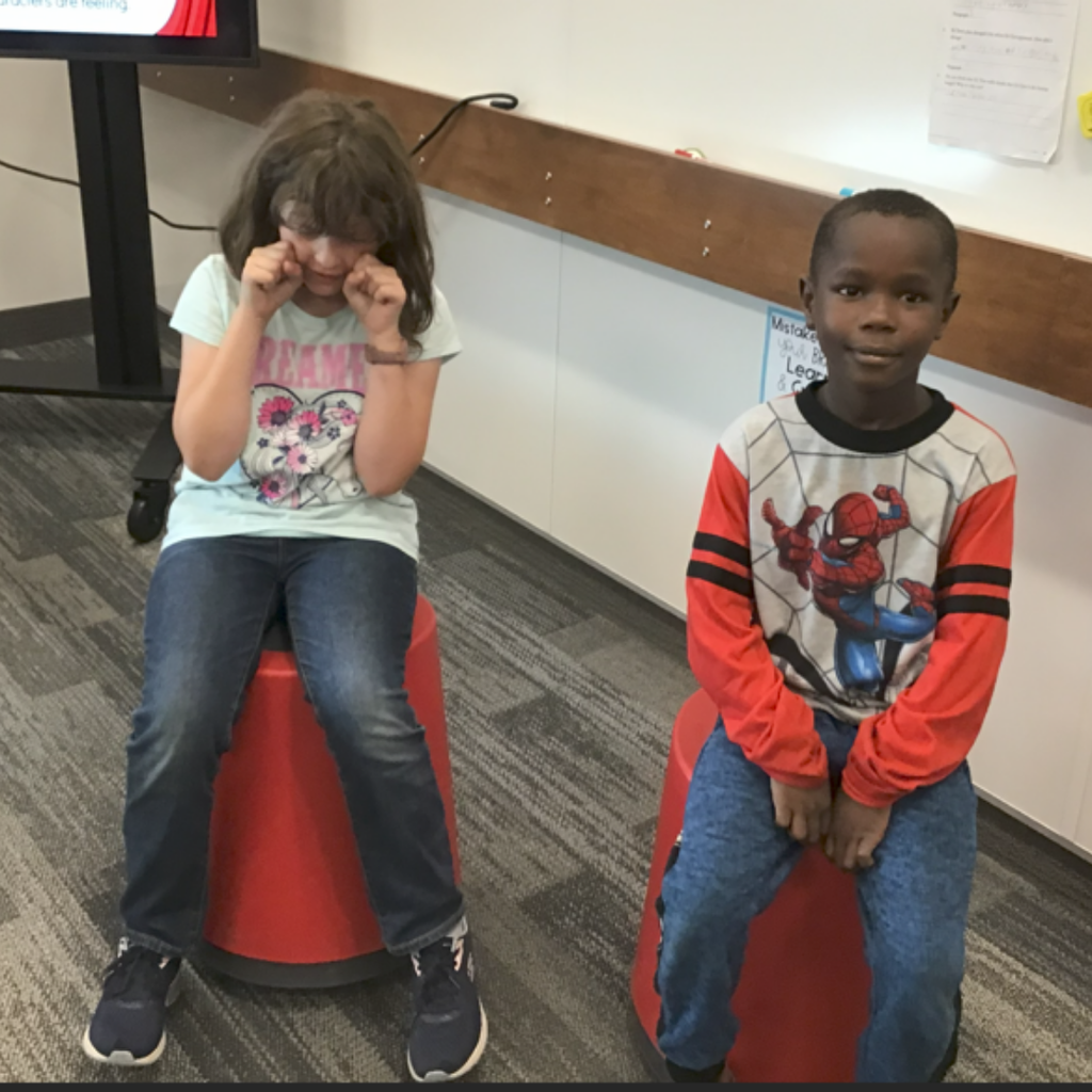 Two students sitting on stools in a classroom. The girl has her head tilted down with her hands balled up near her face, appearing to cry, while the boy sits upright beside her with a calm expression.