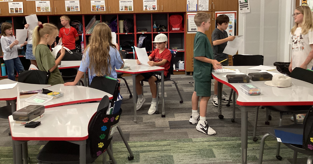 Students in a classroom with some students standing and others sitting. Some students are reading from papers while others are looking at them..