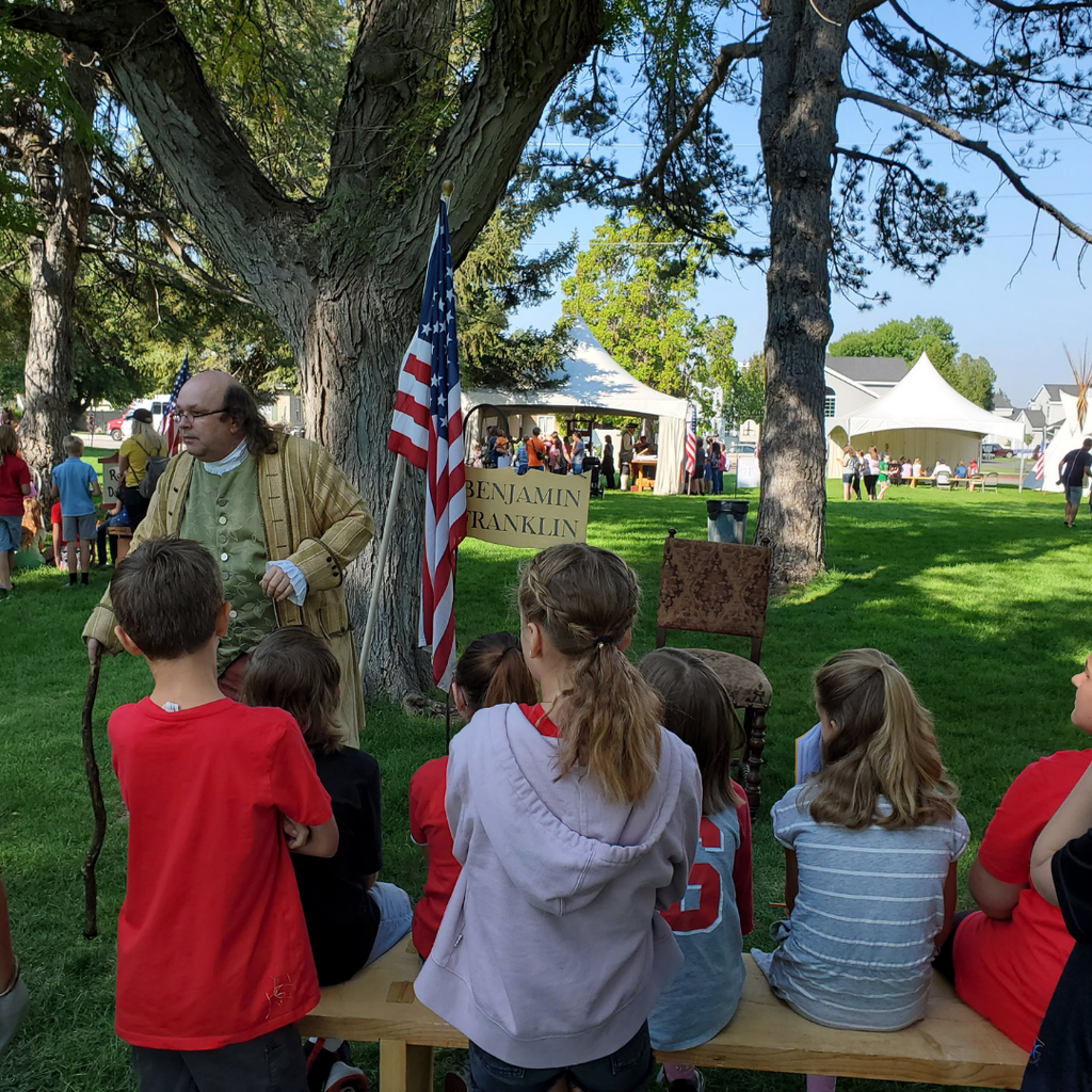 Seven students sit on a bench outdoors, listening to a man dressed as Ben Franklin in 18th-century attire, who is standing next to an American flag.