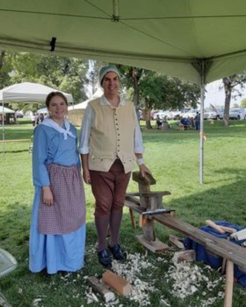 Two people in 18th-century attire stand under an outdoor tent beside a woodworking tool, with wood shavings scattered on the ground.