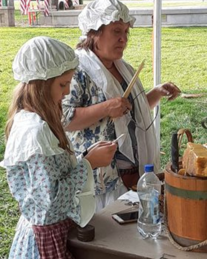 Two women wearing bonnets and dressed in 18th-century costumes, standing outdoors.