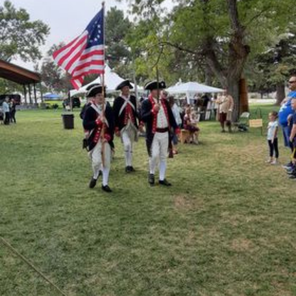 People dressed as 18th-century soldiers marching toward the camera, carrying a colonial-era flag representing the 13 American colonies, while event attendees stand nearby watching.