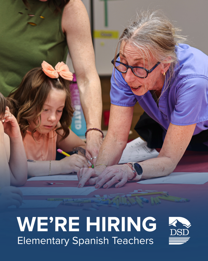 A teacher pointing to something on a paper to help her students. There is a blue banner along that bottom that says: "We're Hiring Elementary Spanish Teachers."
