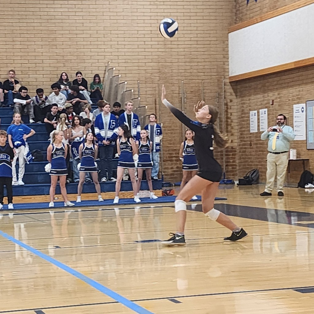 Fairfield's player is serving the ball.  In the background are cheerleaders and Student Body Officers standing with others in the crowd sitting in the bleachers.