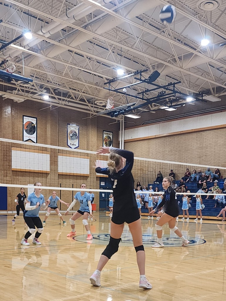 Fairfield's player is preparing to hit a ball over the net.  Another Fairfield player is watching her.  On the other side of the net are Shoreline's players ready for the hit.  In the background are Shoreline's cheerleaders and crowd seated in the bleachers.