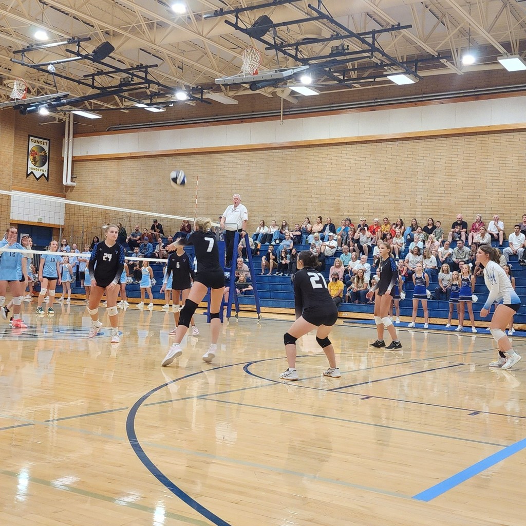 Fairfield's player is passing a ball towards the net while 4 other Fairfield players are watching, ready to play the next pass.  On the other side of the net are Shoreline's players.  In the background the crowd is seated in the bleachers.