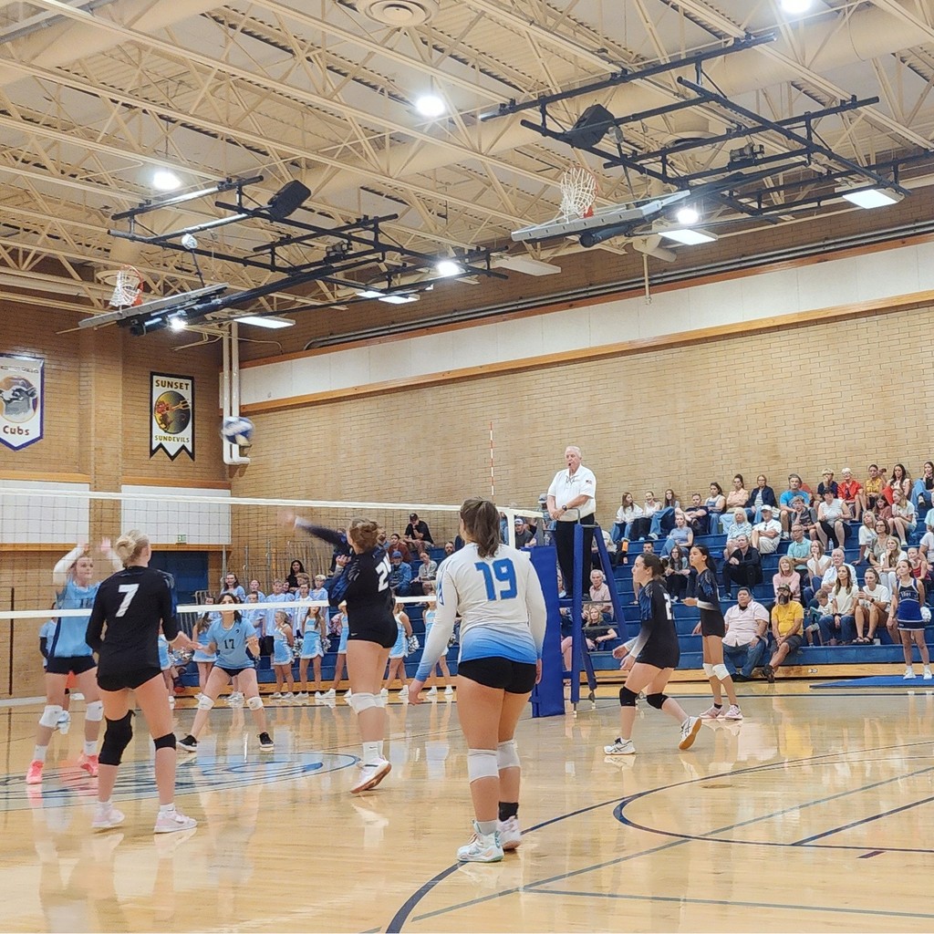 Fairfield's volleyball team is watching one of the Fairfield players hit a ball over the net.  On the other side of the net shoreline is preparing for the hit.  In the background the crowd is seated in the bleachers.