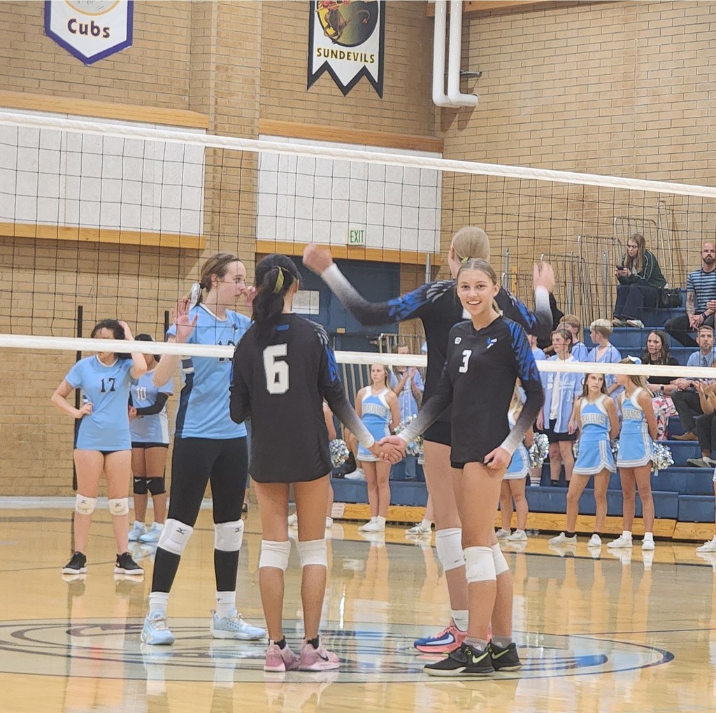 Three of Fairfield's players standing at the net preparing for a serve.  One Fairfield player is looking to the camera and smiling.  On the other side of the net are Shoreline's players, and in the background are Shoreline's cheerleaders  and the crowd seated in the bleachers.