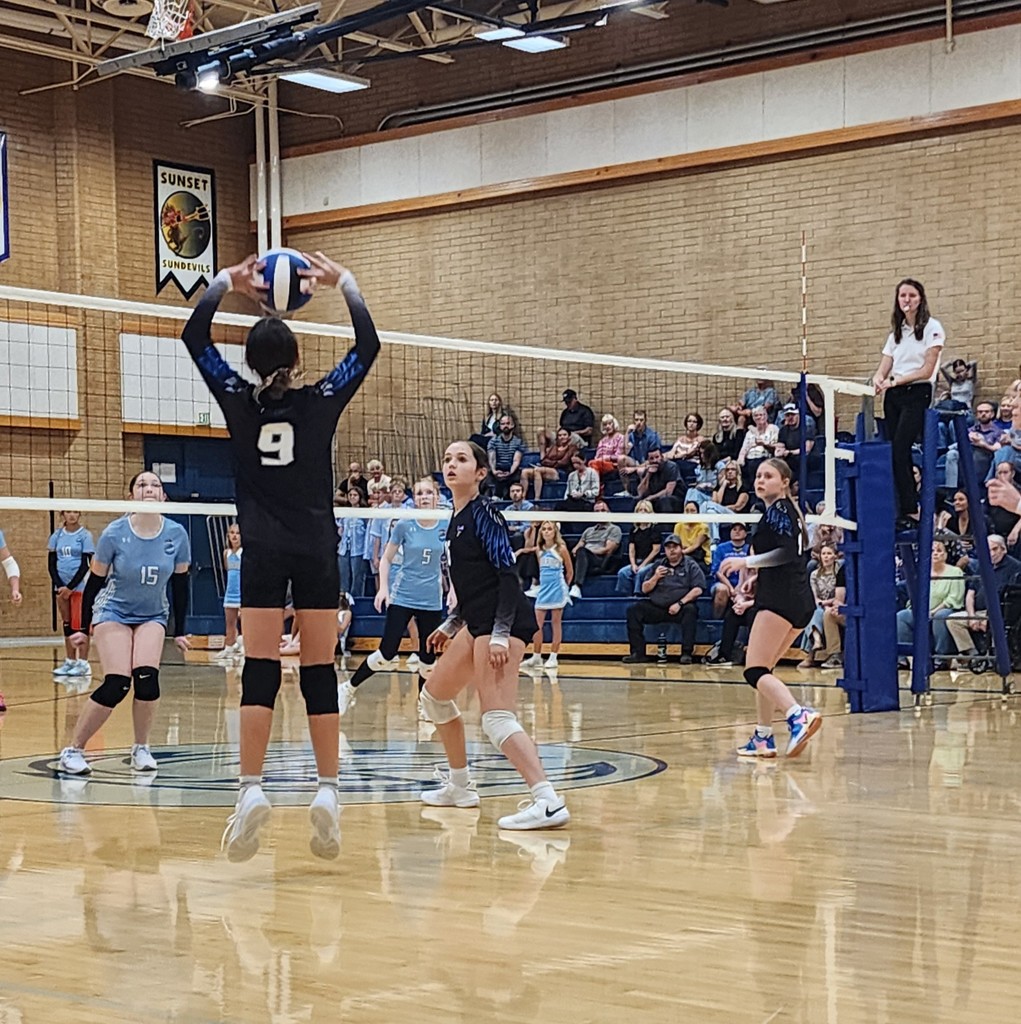 Fairfield's player #9 is setting a ball towards two of Fairfield's front row players who are watching and waiting for the ball.  On the other side of the net are Shoreline's players watching the play.  In the background are crowd members seated in the bleachers.