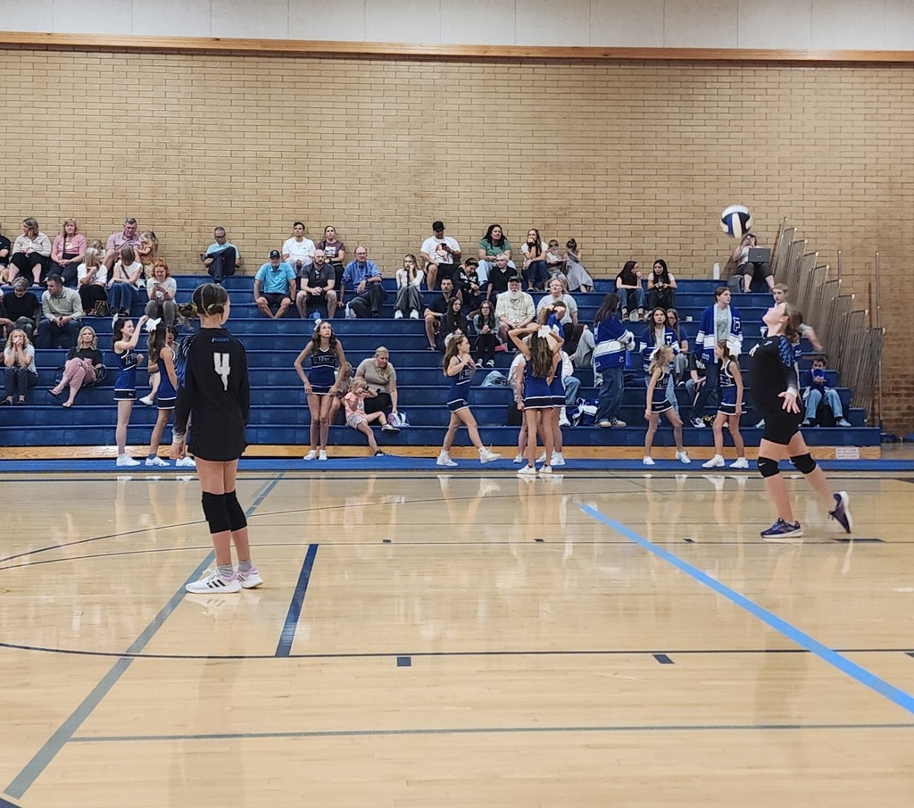 One of Fairfield's JV players is serving the ball.  Another player is watching.  In the background the crowd is seated in the bleachers.