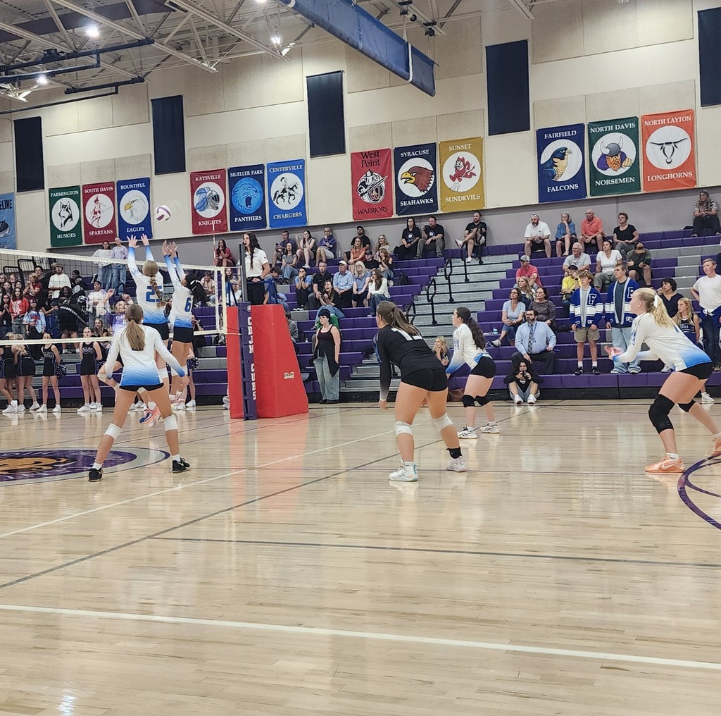 Fairfield is set up to defend an attacked ball.  Two Fairfield players are jumping at the net while the remaining 4 are set to dig the ball.   In the background are the crowd in the bleachers.