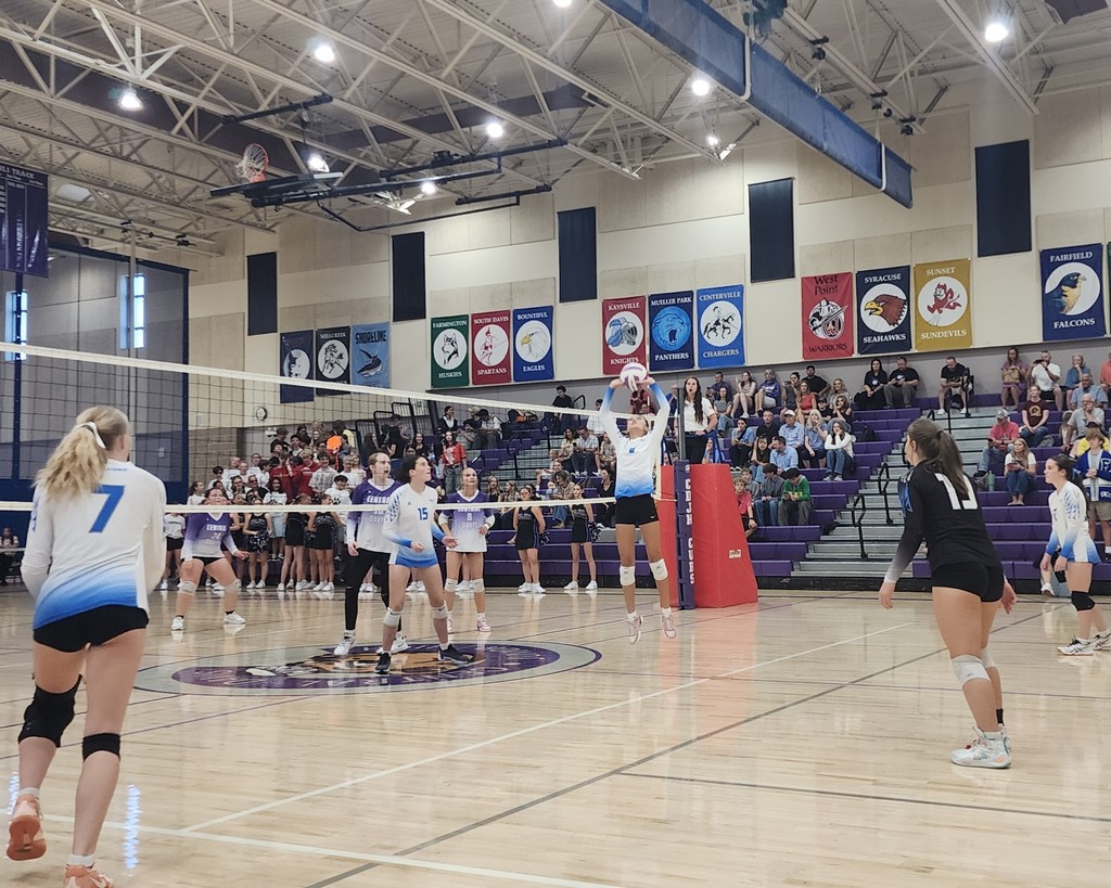 Fairfield's setter is jump-setting the ball to the hitters on our side of the net.  Two players are set up to hit the ball, while a third is watching from the net.  On the other side of the net are Central Davis players watching and preparing.  In the background are crowd-members seated in the bleachers.