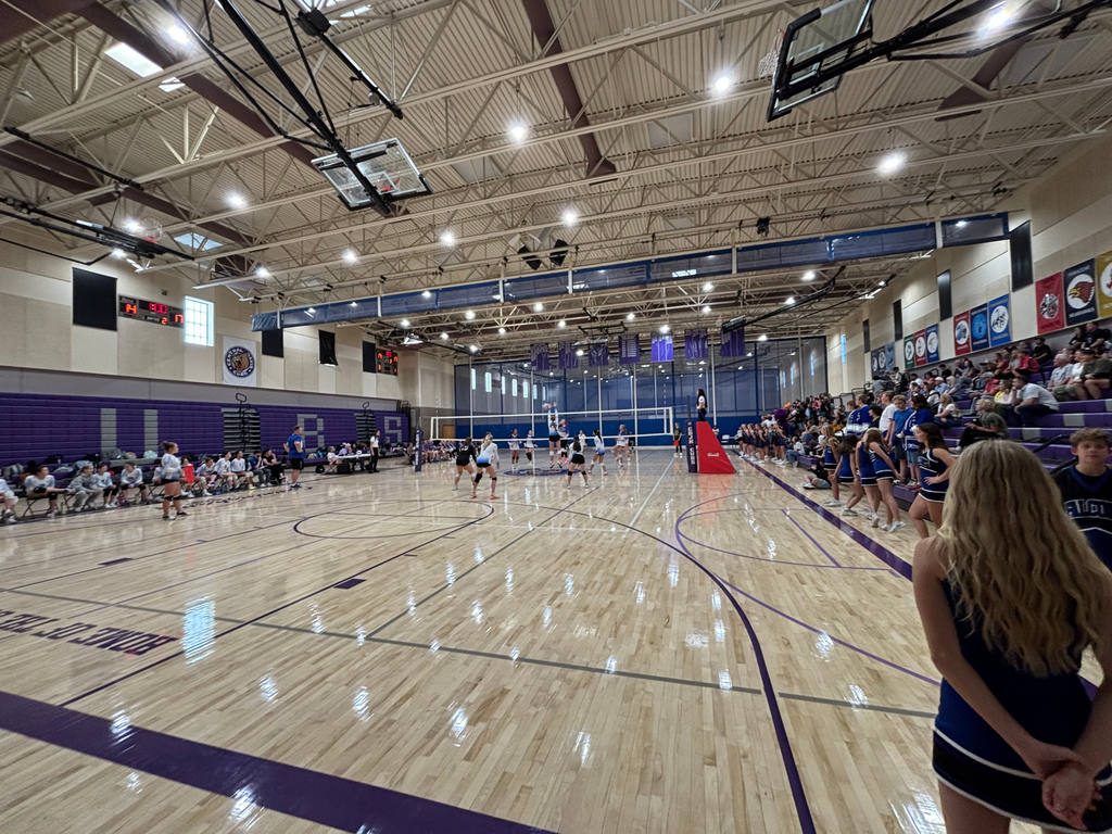 A widescreen image showing the entire gym.  Fairfield's team is on the left, in chairs.  The crowd is on the right in the stands.  Both volleyball teams are in the middle, playing the game.
