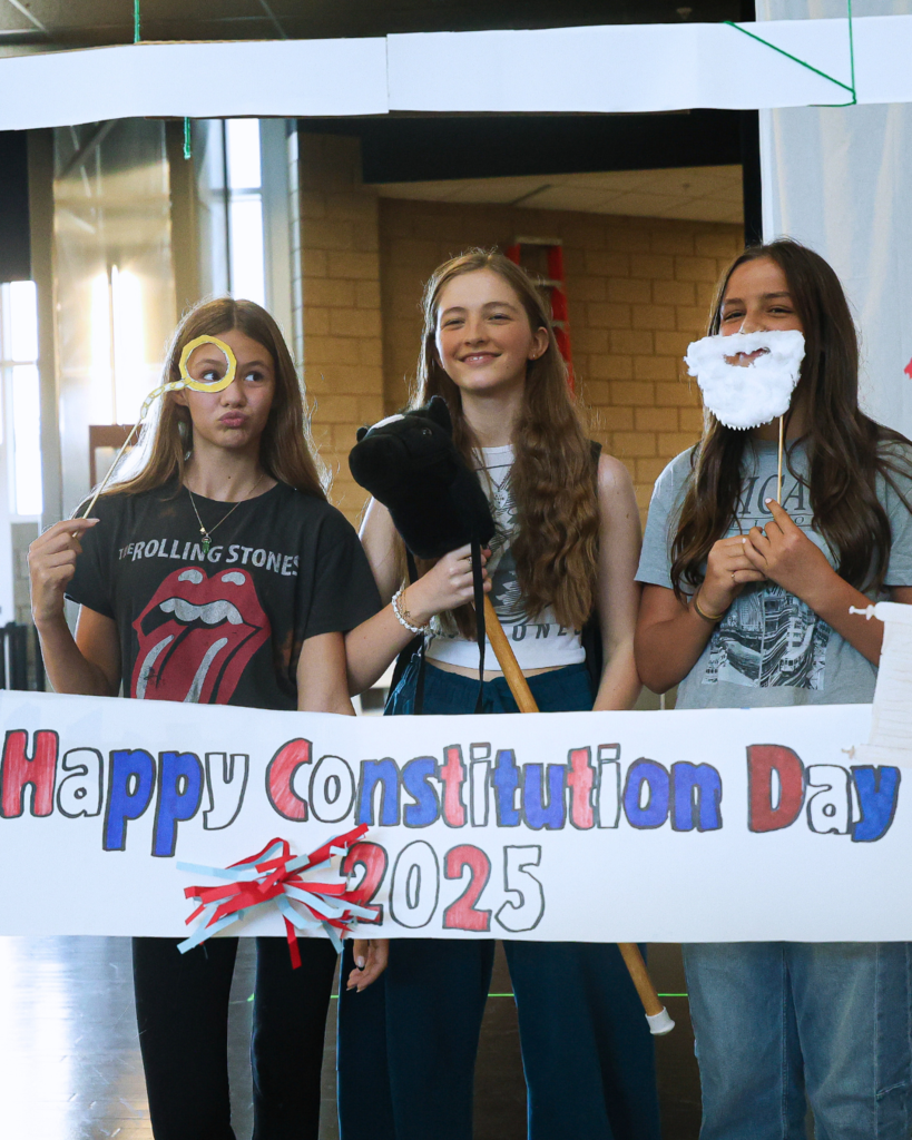 Three students holding up props while standing in front of a "Happy Constitution Day 2025" sign. 