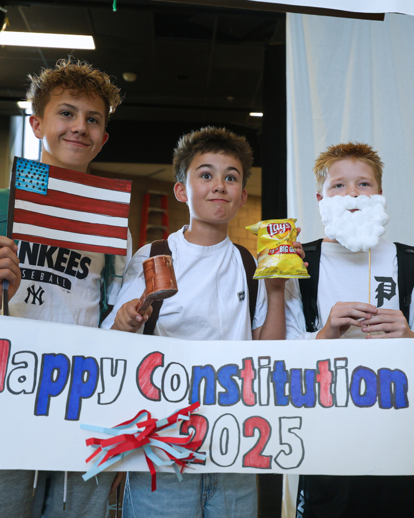 Three students holding up props while standing in front of a "Happy Constitution Day 2025" sign. 
