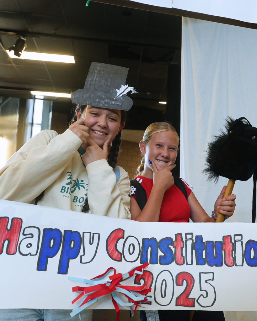 Two students holding up props while standing in front of a "Happy Constitution Day 2025" sign. 
