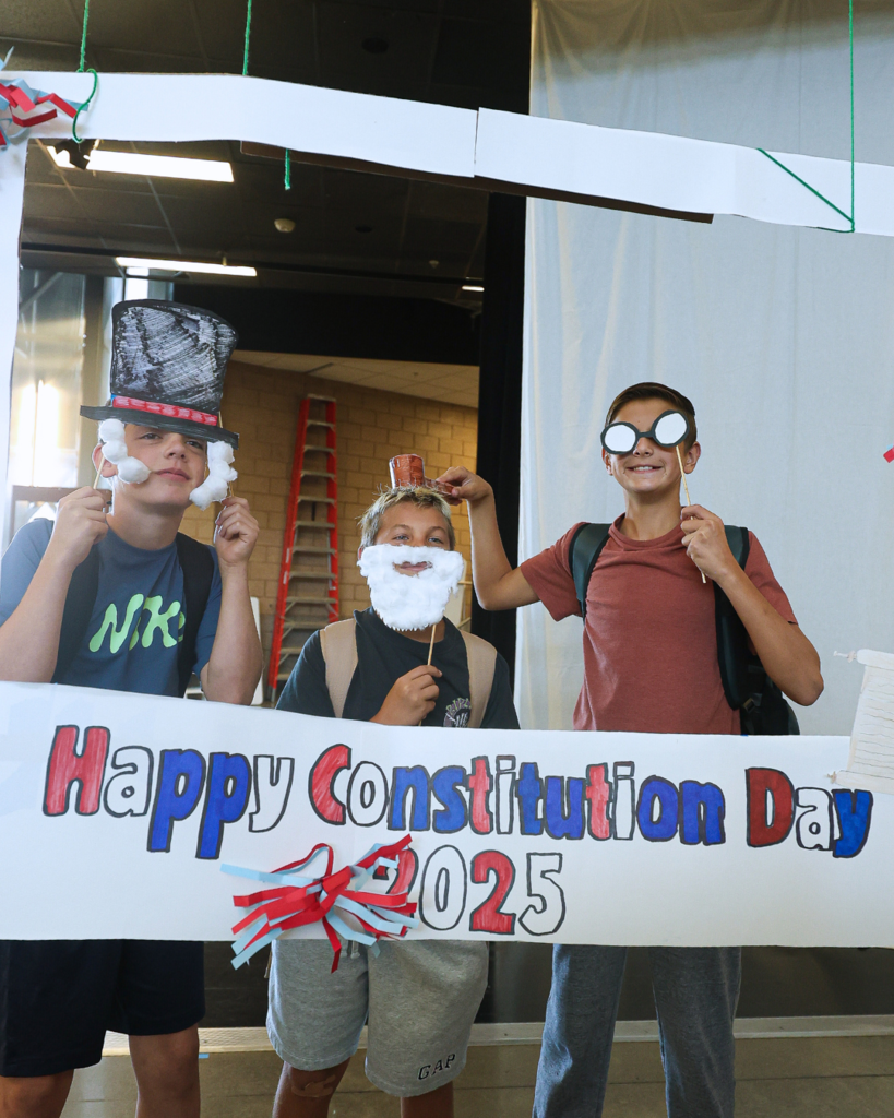 Three students holding up props while standing in front of a "Happy Constitution Day 2025" sign. 