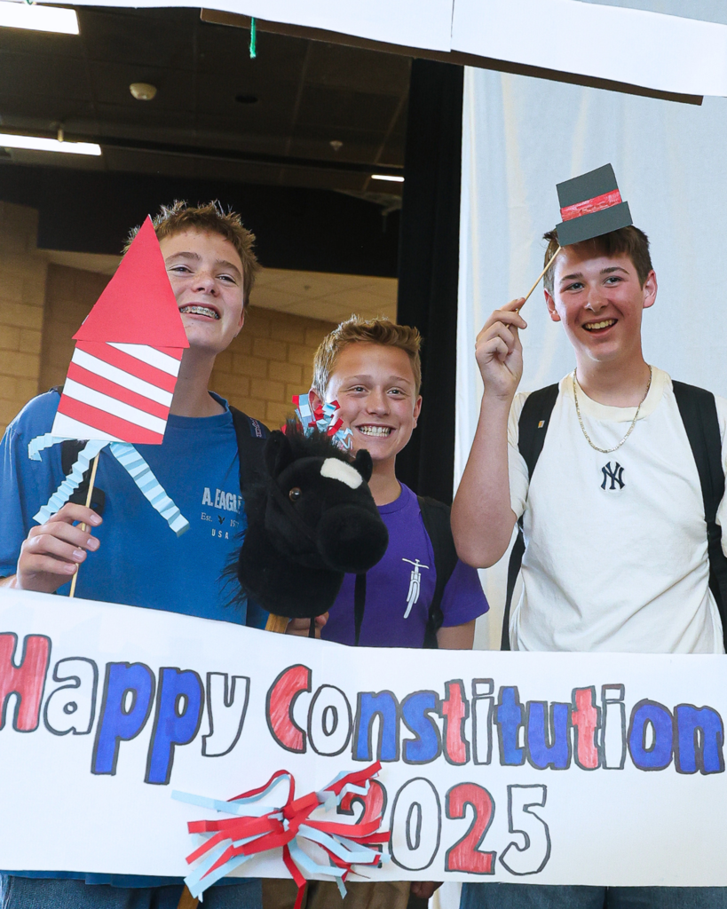 Three students holding up props while standing in front of a "Happy Constitution Day 2025" sign. 