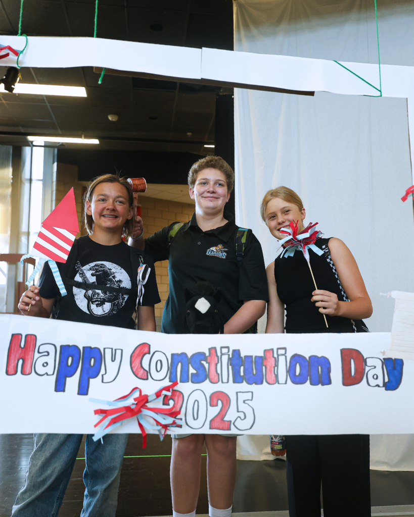 Three students holding up props while standing in front of a "Happy Constitution Day 2025" sign. 