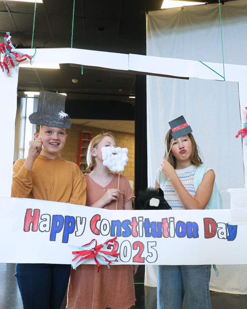 Three students holding up props while standing in front of a "Happy Constitution Day 2025" sign. 
