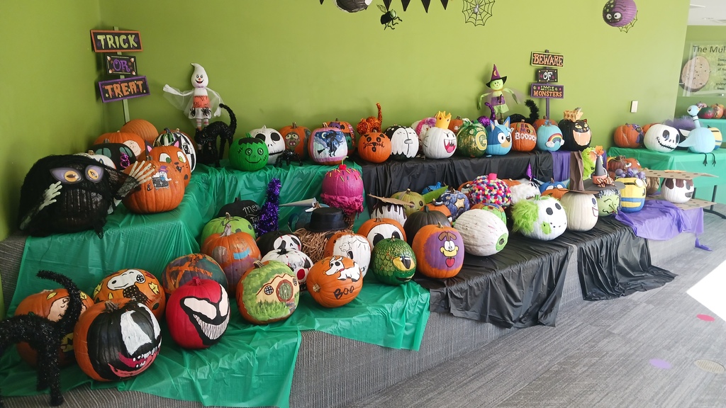 A wide-angle photo showing a collection of decorated pumpkins on display inside a building. The pumpkins are arranged on a series of tiered tables covered with green and purple tablecloths, placed against a bright green wall. Many of the pumpkins are painted with creative designs, including popular characters like the Grinch, Garfield, and an angry bird. Some pumpkins are painted black with white skeleton faces, while others are decorated with glitter, felt, and other craft materials. In the background, Halloween decorations hang from the ceiling, including bats and spiders. A 2 signS on the wall read "BEWARE OF MONSTERS" and "TRICK OR TREAT."