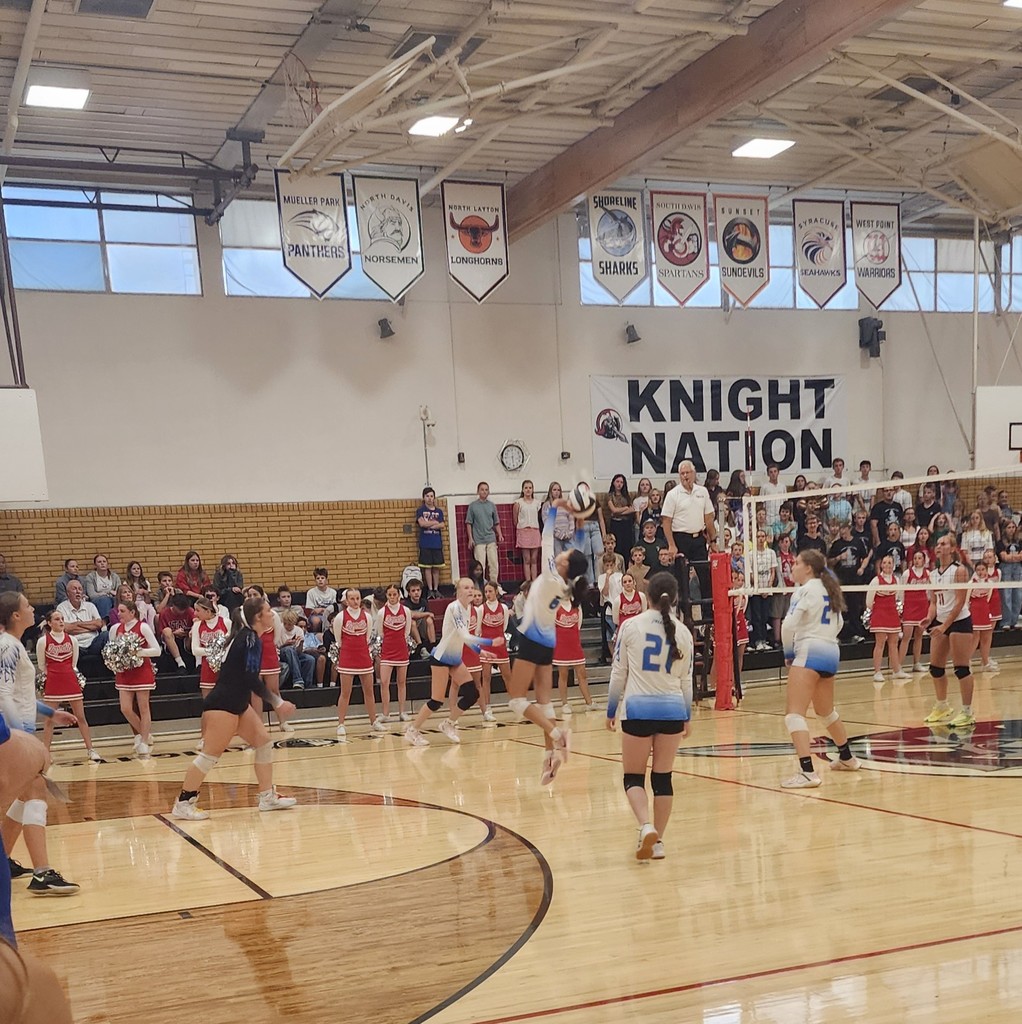 Fairfield's player is setting the ball in the middle of the court.  Towards the left are 2 of Fairfield's players, watching.  Towards her right are 3 of  Fairfield's players moving to hit the ball. 1 of Kaysville's girls are on the other side of the net.  In the background are Kaysville's cheerleaders standing, with the crowd seated and standing behind them in the bleachers.