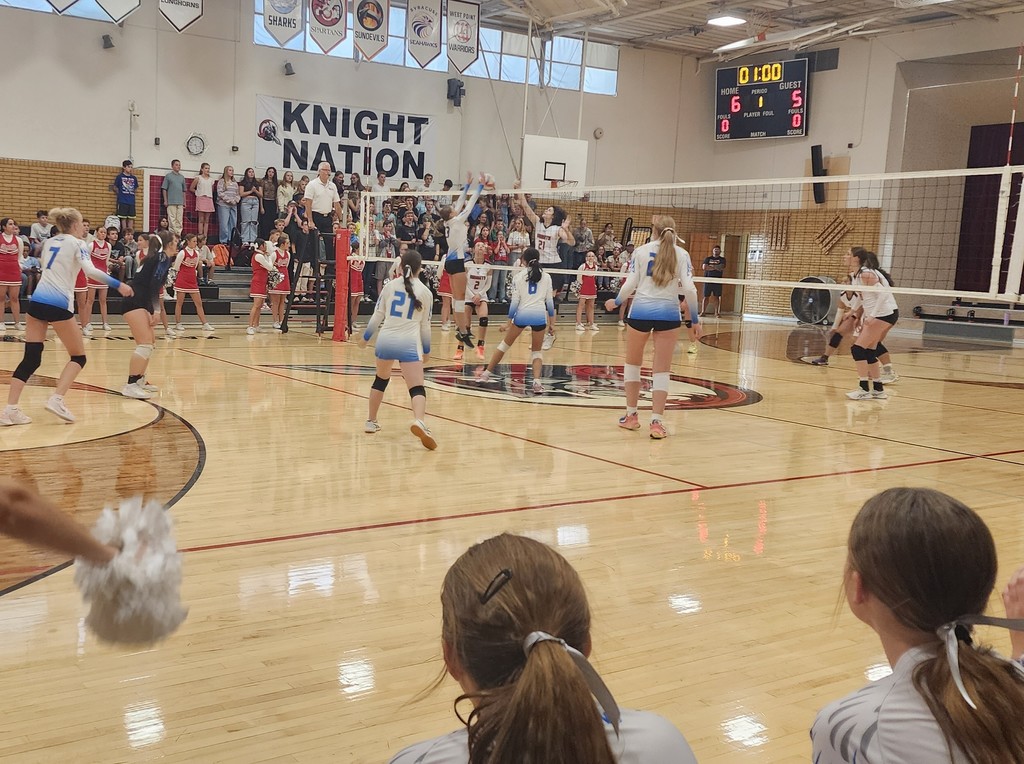 Fairfield's player is jumping to block the ball from coming over the net.  Three other girls are watching from Fairfield's side of the net.  On the other side of the net, Kaysville's player is attacking the ball, while 3 others are watching.  In the fore ground are 2 of Fairfield's players, seated on our bench.  In the background is a crowd of fans sitting and standing in the bleachers.