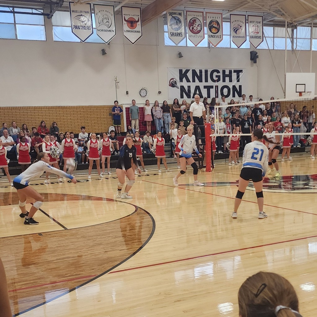 Fairfield's player is passing a ball towards the net.  3 of Fairfield's team are watching, ready to play the next ball.  On the other side of the net is one of Kaysville's players.  In the background are Kaysville's Cheerleaders standing, with the crowd sitting and standing behind them in the bleachers.