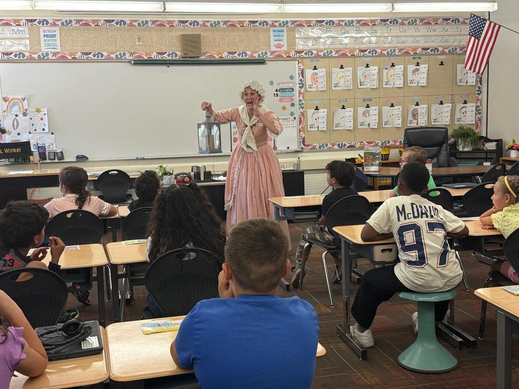 a woman dressed in a historical colonial dress presents to a classroom full of students sitting at their desks. 