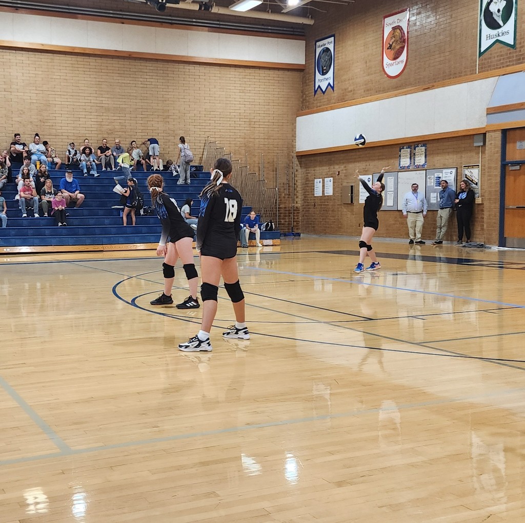 A Fairfield player is serving the ball while two other Fairfield players are watching her.  3 of Fairfield's admin are standing in the back against a wall.  There are several people seated in the bleachers watching the game to the background.