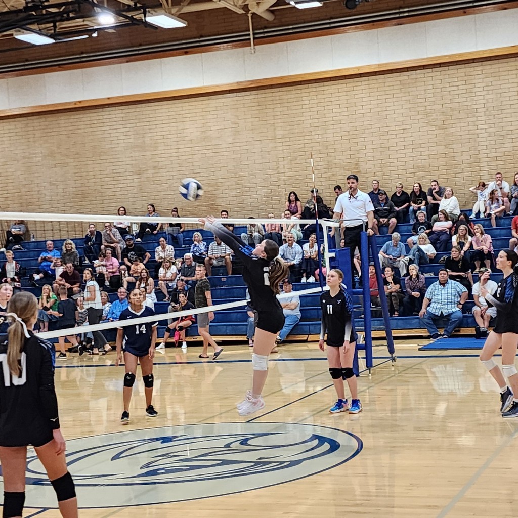 A Fairfield JV player is jumping to block a ball at the net.  Two Fairfield players are watching from the right side, two  Legacy players are to the left side of the net.  The crowd is seated in the bleachers in the background