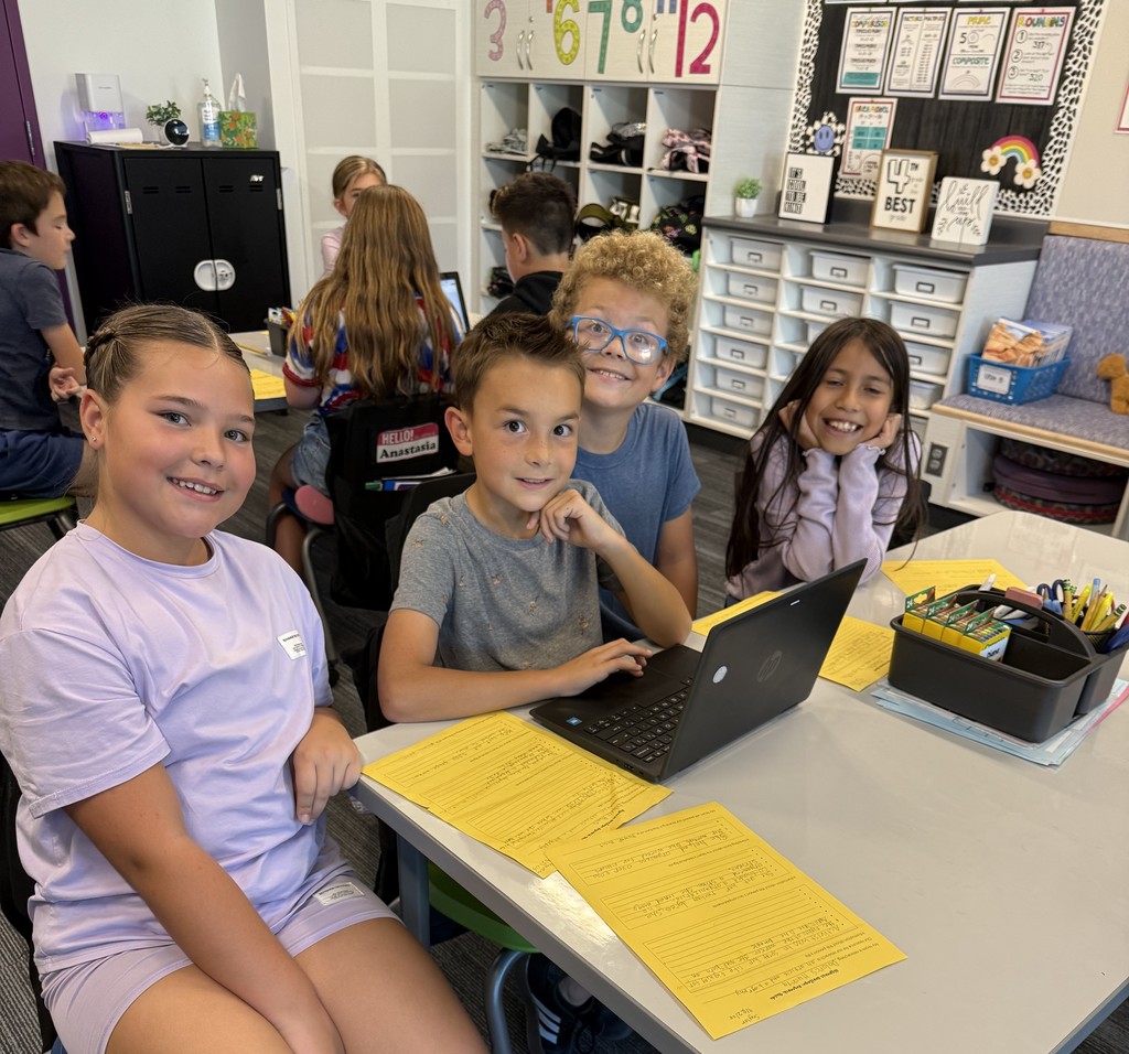 four students smiling sitting in a group with a laptop in front of them and orange papers