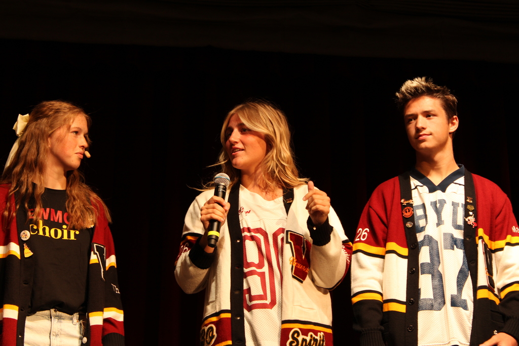 Three Viewmont High students stand on stage in maroon-and-gold letterman cardigans; the center student holds a microphone while the others look on against a dark curtain backdrop.