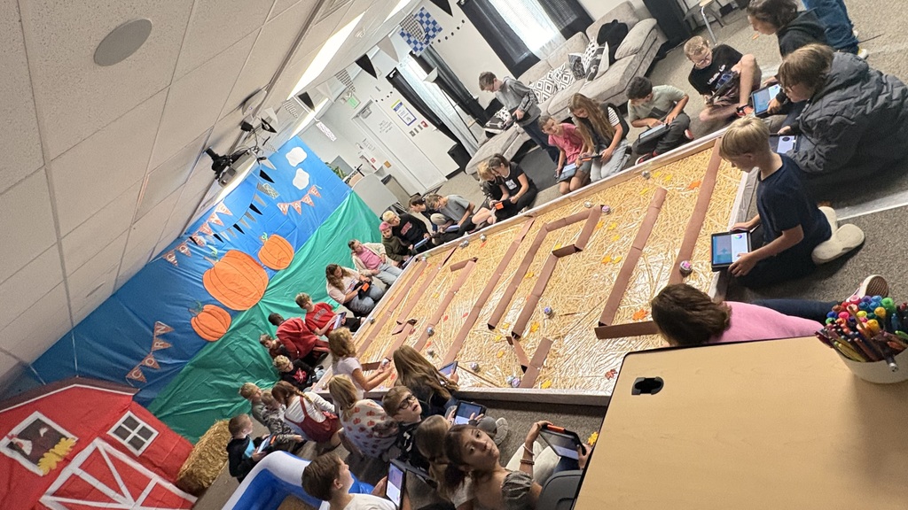 a class sits around the outside of a maze with ipads and robots with a barnyard background