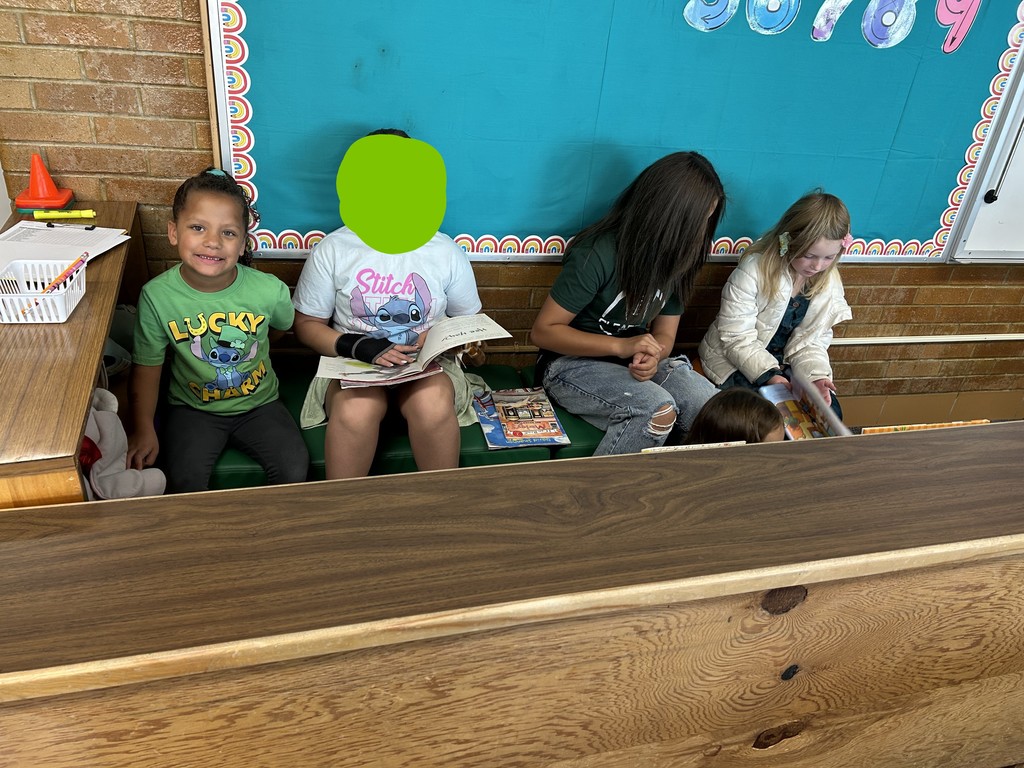 Two older students and two younger students sit side-by-side on a wooden bench seat, with a brick wall and a teal bulletin board behind them. The students are looking at books during their reading buddy time. The younger student on the far left is smiling at the camera.